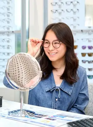 A woman is smiling and looking at herself in a mirror while wearing glasses and sitting in front of a desk with a keyboard and other objects.