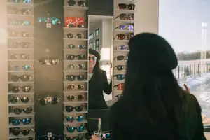 A woman wearing a hat and sunglasses is looking at herself in a mirror in a store that displays various sunglasses.