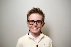 A smiling young boy with glasses and a light beige jacket is posing for a photo against a white background.