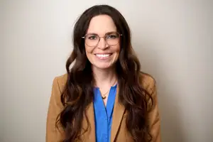 A woman with long brown hair wearing a blue top, a brown blazer, and eyeglasses, smiling and standing in front of a white wall