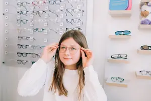 A young girl trying on glasses in an optical shop with a display of different frames behind her.