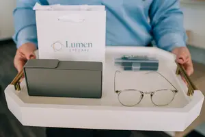 A person is holding a white tray with a pair of glasses and a box with the text 'Lumen Eyecare'.