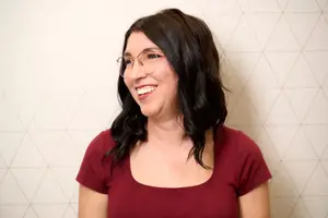 A woman with glasses smiling in front of a white wall with a geometric pattern