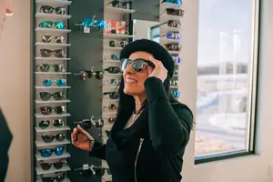 A woman in a black outfit is standing in front of a wall of sunglasses, smiling, and adjusting her sunglasses.