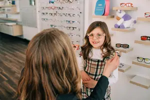 A girl in a plaid dress is trying on glasses in a store with a woman helping her.