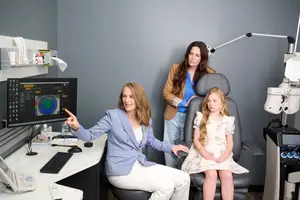 Three women are in an eye care clinic, with one woman seated and pointing at a monitor, another standing behind, and a young girl sitting in a chair looking at the woman pointing.