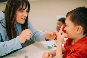 A woman is adjusting a young boy's eyeglass. Behind them, another person is looking at a mirror.