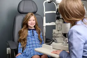 A young girl in a blue dress is sitting in a chair and smiling at the camera while an eye doctor checks her eyes using an ophthalmoscope.