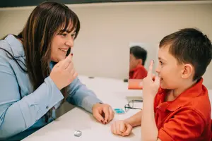 A woman and a young boy are seated at a table, with the woman showing something to the boy and both of them smiling, and a mirror reflecting another person behind them.
