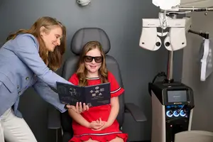 A woman is helping a girl wear sunglasses and looking at a book with the text '- - E', and the girl is wearing a red dress and smiling. A machine is placed behind the woman. The background is gray.