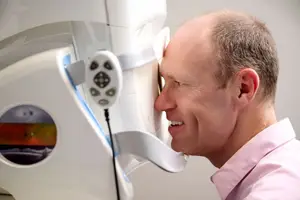 A man wearing a pink shirt is looking into a machine with a screen displaying an image of a beach and a mountain range