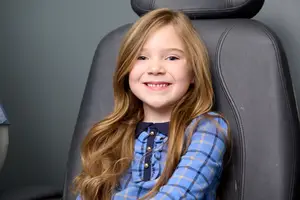A young girl with long blonde hair is smiling and sitting on a black leather chair in a room with gray walls.