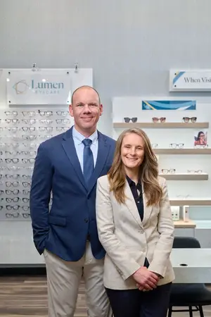 A man and a woman standing together in an office room with a table, chairs, and shelves with glasses, and a sign that reads 'Lumen Eyecare'.