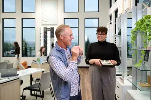 A man and a woman are standing inside a room with wooden floors and large windows. The man is wearing glasses and a blue vest, while the woman is holding a box and wearing glasses and a black turtleneck. There are several people walking in the distance, and there are chairs, a table with a laptop, a plant, and a shelf with a plant in the room.