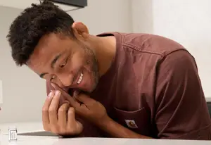 A man is smiling and looking at something while he is holding a glass cup and biting his teeth in the kitchen area.
