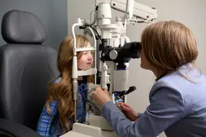 A young girl with long blonde hair is sitting in an ophthalmologist's chair while an adult woman is adjusting a device in front of her.
