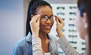 A woman is trying on glasses while a person holds them in front of her face, and they are both smiling at each other in a room with a wall full of glasses and a blurry background.