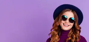 A woman in a purple turtleneck and sunglasses smiling standing against a purple background