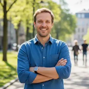 A smiling man with short brown hair is standing with his arms crossed on a sunny day, with people and buildings in the background.