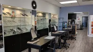 A photo of an indoor optical shop with several glass cabinets, tables, chairs, and a black wall with a mirror, as well as a poster on the right side.