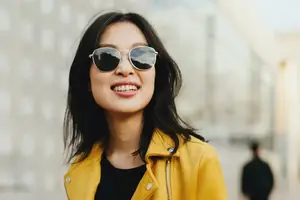 A woman wearing a yellow jacket, black top, and silver sunglasses is smiling and standing in an outdoor area, with a blurry background of a person walking and a building behind her.