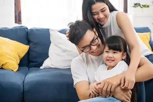 A family sitting on a couch, with a man wearing glasses and a woman wearing a sleeveless top holding a child, all smiling at the camera.