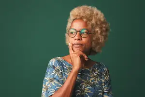 A woman with curly hair wearing glasses and a blue shirt with palm trees on it stands against a green background.