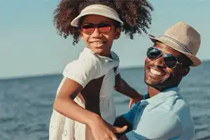 A man and a young girl are smiling at the camera while wearing hats and sunglasses. They are standing on the beach near the water, with the man holding the girl's hands.