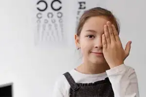 A young girl covering her eyes with her hand in a room with an eye chart on the wall