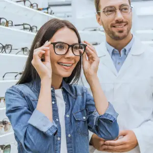 A smiling woman trying on glasses with a man in a white lab coat behind her in an optometrist's office