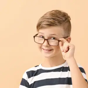 A boy with brown hair wearing a striped shirt and eyeglasses is smiling and posing for a picture with his left hand on his face.