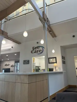 A reception desk in a medical office with a sign that reads Eye Care Professionals, surrounded by lamps and a framed certificate, a monitor on the counter, and a lamp and couch in the background.