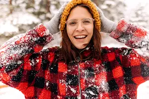 Woman wearing red and black plaid coat, yellow knitted hat, and gray gloves with snow on them smiling in the snow