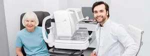A man and an elderly woman are sitting in front of an eye-testing machine. The woman is wearing a blue t-shirt, and the man is wearing a white shirt and a white coat. They are both smiling and looking at the camera.