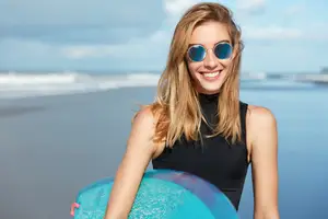 A woman is smiling and holding a surfboard on the beach.