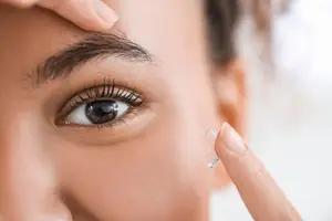 Close up of a woman's face with one eye visible and her hand holding a contact lens in front of her eye