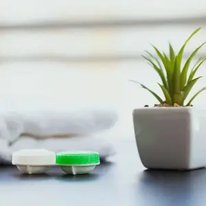 A plant with green leaves in a white pot next to two white and green contact lens containers on a table with a white towel in the background.