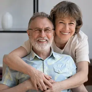 An elderly man wearing glasses and a woman with short hair are smiling and posing for a photo with their arms around each other