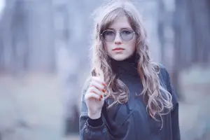 A girl with long blonde hair and red nail polish stands in a forest, wearing a black jacket and glasses.