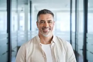 A smiling man in a white shirt stands in an office hallway with glass walls and doors.