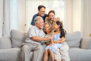A family is sitting on a couch, smiling and hugging each other in a living room with a white curtain in the background.