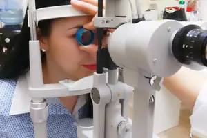 woman with long black hair wearing a white uniform with blue floral print and a silver earring getting her eye examined with a medical ophthalmoscope
