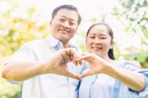 A couple is posing for a picture with their hands shaped like a heart, smiling and standing near a tree in a park during the daytime