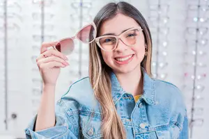 A woman with long blonde hair is wearing a denim jacket and holding a pair of pink glasses.