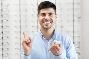 A man in a blue shirt holds up a lens in front of a wall of glasses.