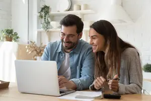 A couple sits at a table, with a woman smiling at the man who is looking at a laptop.