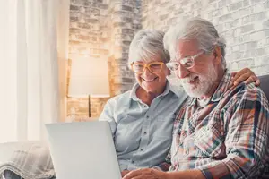 A senior couple sits on a couch and uses a laptop, with a brick wall in the background.