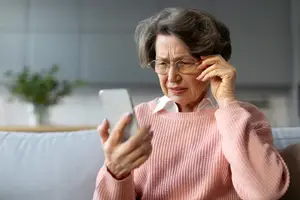 An elderly woman wearing glasses and a pink sweater is sitting on a couch and looking at her phone.