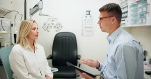 A woman and a man are inside a room that looks like an optometrist's office. The man is speaking to the woman, and she is listening attentively. Behind them is a wall with an eye chart and a black chair. The man is wearing glasses and a watch, while the woman is wearing a white shirt and a bracelet.