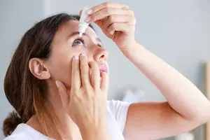 A woman is applying eye drops to her eye in front of a mirror.